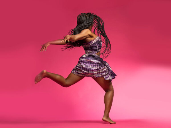 Beautiful African Black girl wearing traditional colorful African outfit does a dramatic dance move against a colorful pink background
